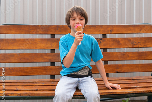 The boy is sitting on a bench and enjoys eating pink ice cream. © got