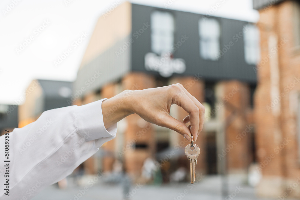 House keys in female hand on background of new buildings and sky. Real estate agent, moving home or renting property. Woman hand holds the keys to an apartment against the background of buildings.