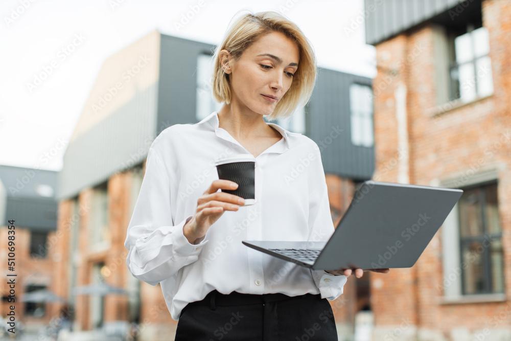 Attractive young businesswoman in white shirt working on laptop. Professional business woman using modern digital device smiling drinking coffee near office building in city. Attractive young businesswoman in white shirt working on laptop. Professional business woman using modern digital device smiling drinking coffee near office building in city.