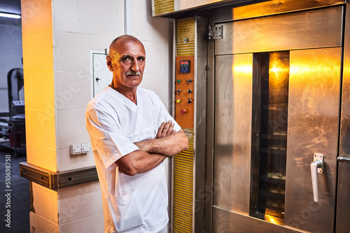 Portrait of professional baker in uniform stands on the background of industrial oven in a bakery for making bread