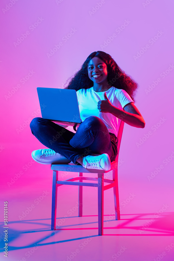 Full length of young black lady showing thumb up, using laptop, recommending remote studies or online work in neon light Full length of young black lady showing thumb up, using laptop, recommending remote studies or online work in neon light