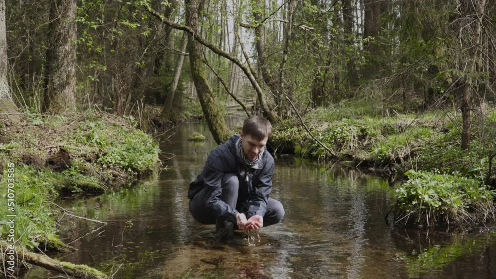 Happy man on hike squats in rubber boots in shallow transparent river among wild forest. Tourist picks up healthy clear cool water in handful, drinks it. Traveler enjoys rest, scenery and smiles. Happy man on hike squats in rubber boots in shallow transparent river among wild forest. Tourist picks up healthy clear cool water in handful, drinks it. Traveler enjoys rest, scenery and smiles.