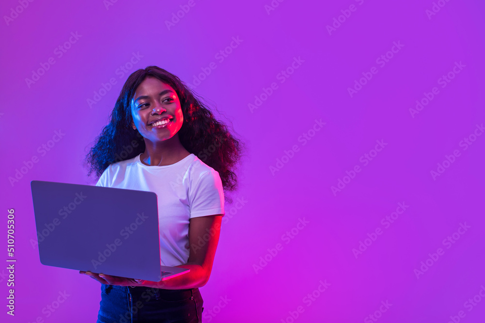 African American lady using laptop for online work, education or communication, looking at empty space in neon light - obrazy, fototapety, plakaty 