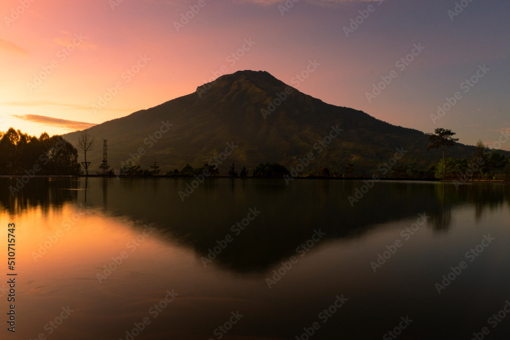 Sunrise with Mount Sumbing with lake surface on the foreground. The lake surface make reflection of mountain and sunrise sky. Embung Kledung, Central Java, Indonesia
