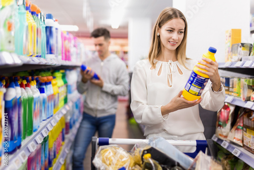Portrait of young smiling cheerful positive woman with shopping cart choosing household chemicals in supermarket