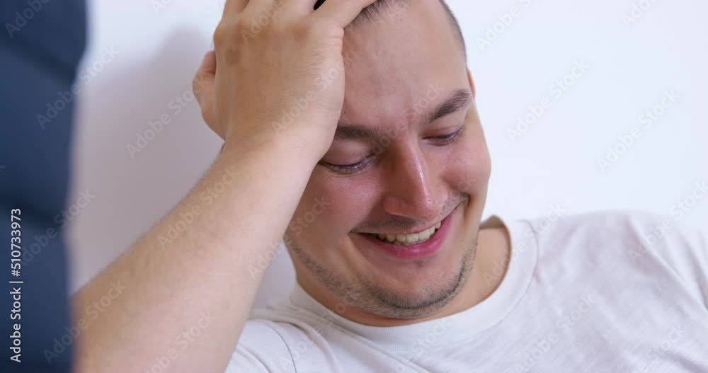 Young man laughs and talks putting hand on head at home. Guy sits against white wall watching and discussing videos in smartphone closeup Young man laughs and talks putting hand on head at home. Guy sits against white wall watching and discussing videos in smartphone closeup