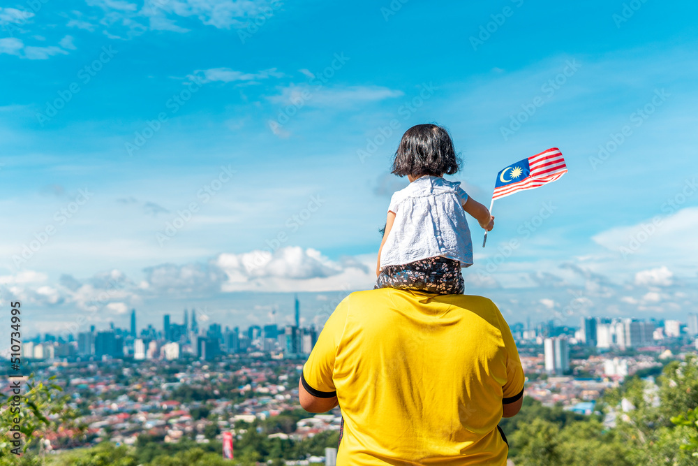 Fototapeta premium Local father and daughter holding a Malaysian flag at the top of a hill. All over Malaysia will be flagged for the celebration of independence day in month of august.