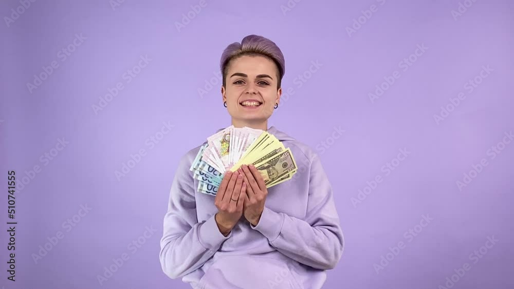 Joyful young adult woman generation Z with short purple hair holding bunch of different currencies, tossing them up, rejoicing, boasting money profit. Indoor studio shot isolated on purple background Joyful young adult woman generation Z with short purple hair holding bunch of different currencies, tossing them up, rejoicing, boasting money profit. Indoor studio shot isolated on purple background