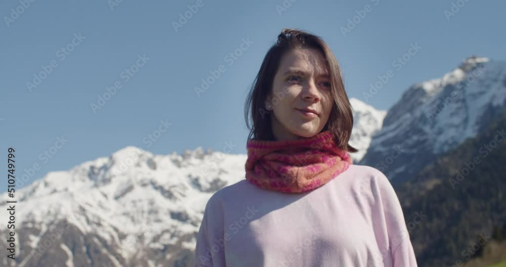 Portrait of young woman looking at camera and smiling on background of mountains. Tourist girl standing outdoors alone enjoying beautiful moment in nature. Happy tourist during travel in rays of sun Portrait of young woman looking at camera and smiling on background of mountains. Tourist girl standing outdoors alone enjoying beautiful moment in nature. Happy tourist during travel in rays of sun