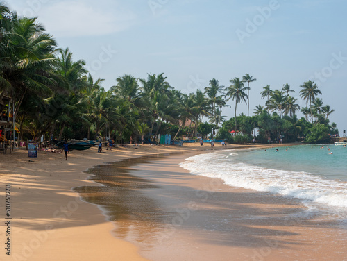 Hikkaduwa, Sri Lanka - March 4, 2022: Beautiful view of the beach in Hikkaduwa with bright green palm trees. People relax and swim in the Indian Ocean
