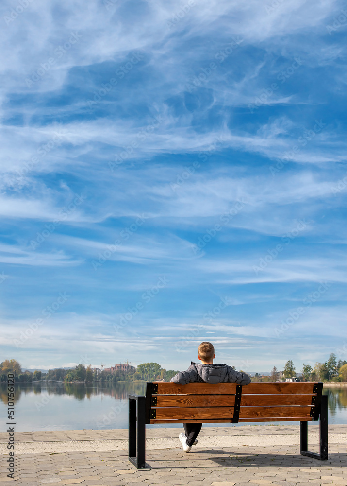 White clouds in the sky. A young guy sits on a bench by the lake. Feeling Blue. Towards the future. Disease prevention. World mental health day White clouds in the sky. A young guy sits on a bench by the lake. Feeling Blue. Towards the future. Disease prevention. World mental health day