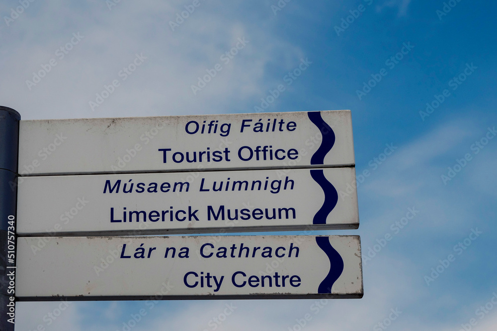 Sign in English and Irish language in Limerick city with direction to Tourist office, Museum and city centre. Blue cloudy sky background.