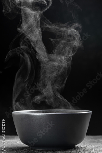 Bowl of hot steam of hot soup with smoke. black ceramic bowl on dark background. Hot food. Culinary, cooking, concept
