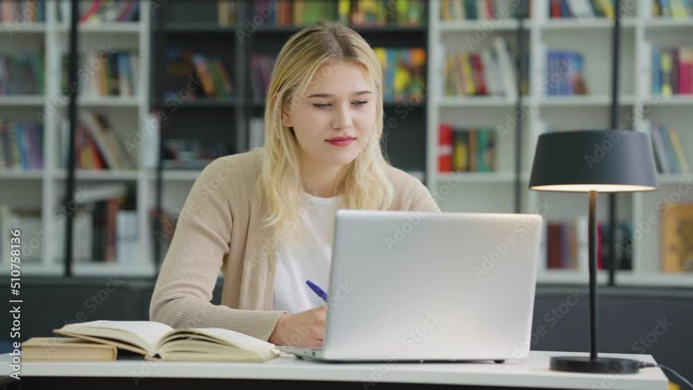 Teenage girl with long blonde hair sitting at table and doing research using laptop reading books. Student doing assignment in library, bookshelves on background Teenage girl with long blonde hair sitting at table and doing research using laptop reading books. Student doing assignment in library, bookshelves on background
