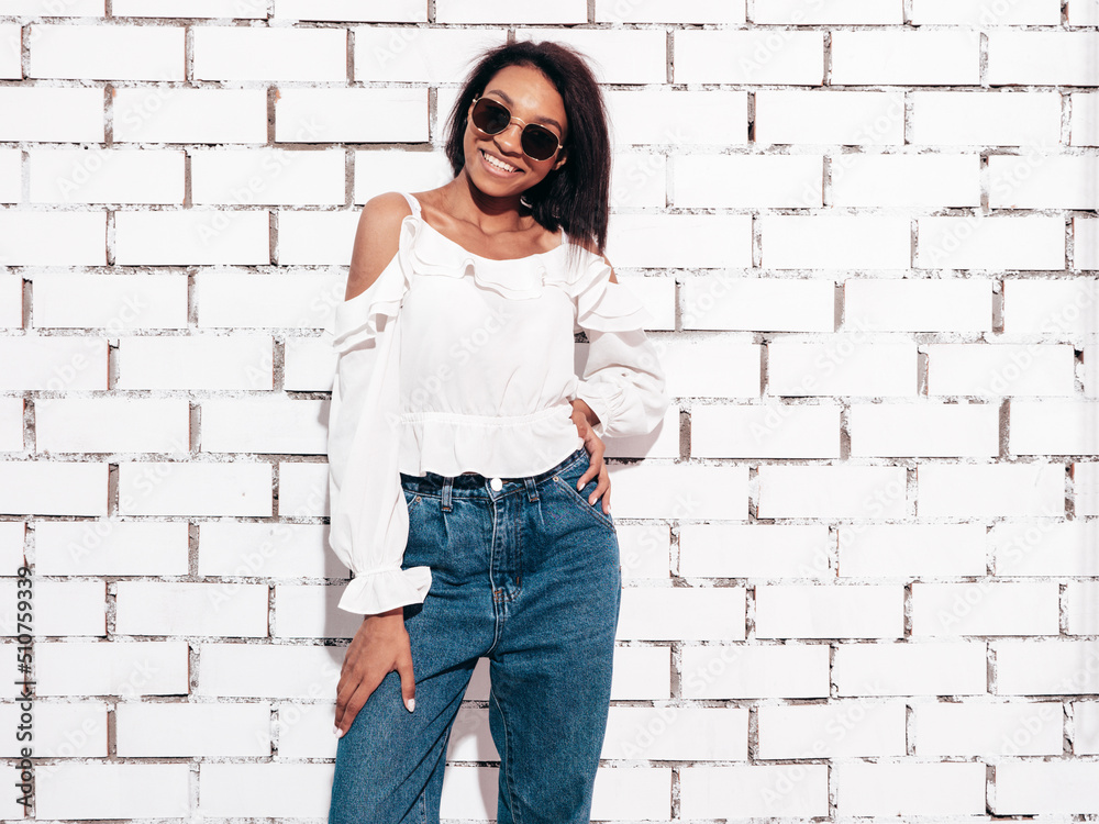 Portrait of young beautiful black woman. Smiling model dressed in summer jeans clothes. Sexy carefree female posing near white brick wall in studio. Tanned and cheerful. In sunglasses