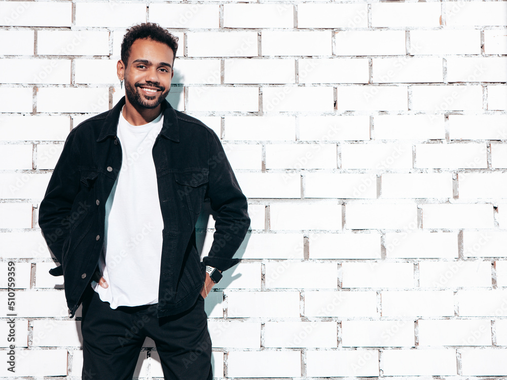 Handsome smiling hipster model. Sexy unshaven man dressed in black summer shirt and jeans clothes. Fashion male posing near white brick wall in studio at sunny day. Isolated Handsome smiling hipster model. Sexy unshaven man dressed in black summer shirt and jeans clothes. Fashion male posing near white brick wall in studio at sunny day. Isolated