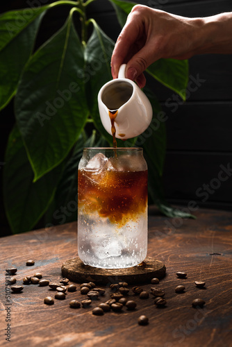 Espresso tonic in the making. Adding coffee into a highball glass filled with ice cubes and tonic soda water, coffee beans, milk jug, wooden background