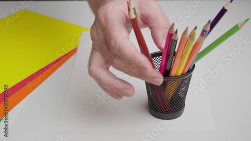 The artist puts colored pencils in a stand. A glass of colored pencils on a white table with drawing paper.