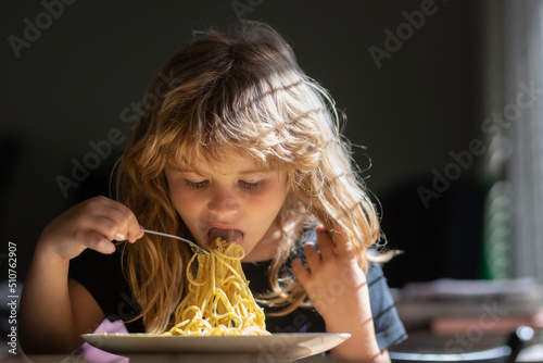 Cute child eats food itself with spoon. Cute little kid eating spaghetti pasta at home.