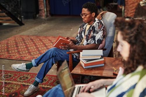 Serious African American guy in stylish shirt and blue jeans opening book in brown cover while sitting in armchair in living room at leisure