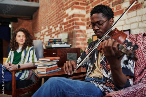 Young man in eyeglases and casualwear sitting in armchair in living room of loft apartment at leisure and playing violin for his girlfriend photo