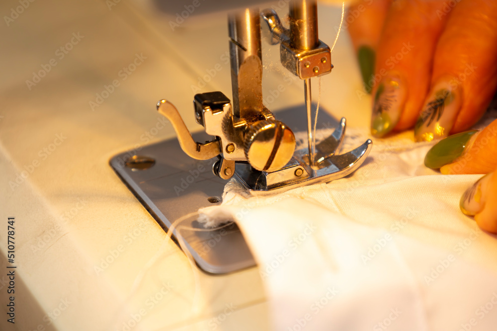 Close up view of sewing process. Female hands stitching white fabric on professional manufacturing machine at workplace. Seamstress hands holding textile for dress production. Light blurred background