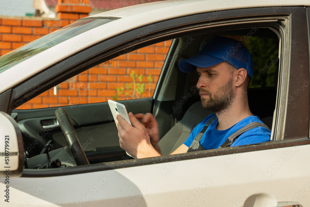 Naklejka premium Bearded man in blue cap sits on driver seat and makes notes about internal car details on tablet. Auto mechanic types on tablet diagnosing car state closeup