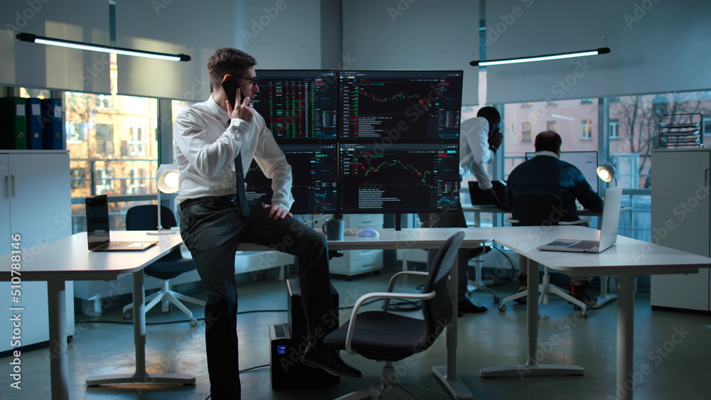 Businessman talk on smartphone sitting at desk with multiple screens with financial data