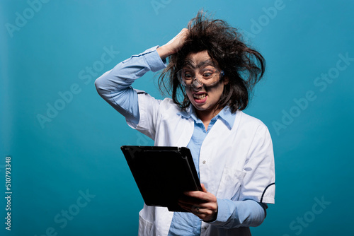 Mad angry biochemist investigating failed experiment report on tablet while pulling hair like a crazy person. Enraged lunatic scientist with touchscreen device, dirty face on blue background.