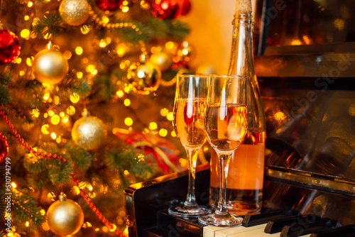 Bottle of white wine and two wineglasses full of drink on the piano with background of Christmas tree. Celebrating Christmas or New Year with a partner, romance concept