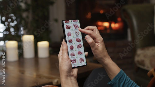 Ordering food using a smartphone at home. A woman selects meat and fish in an online store using an application on a smartphone. Home evening furnishings with a burning fire in the fireplace.