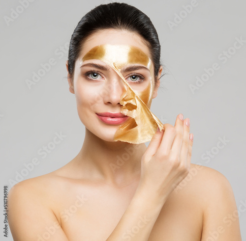 Beauty Woman peeling off Golden Facial Mask. Smooth Skin Model taking off Gold Purifying Face Film Mask over Gray. Women cleansing Cosmetics and Skincare Cosmetology photo