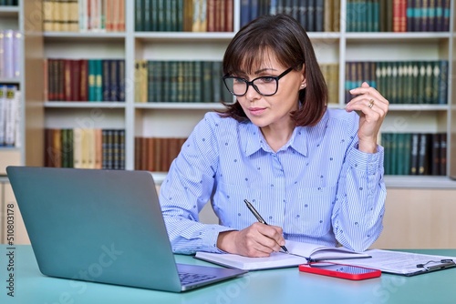 Middle-aged female teacher working in library, using laptop