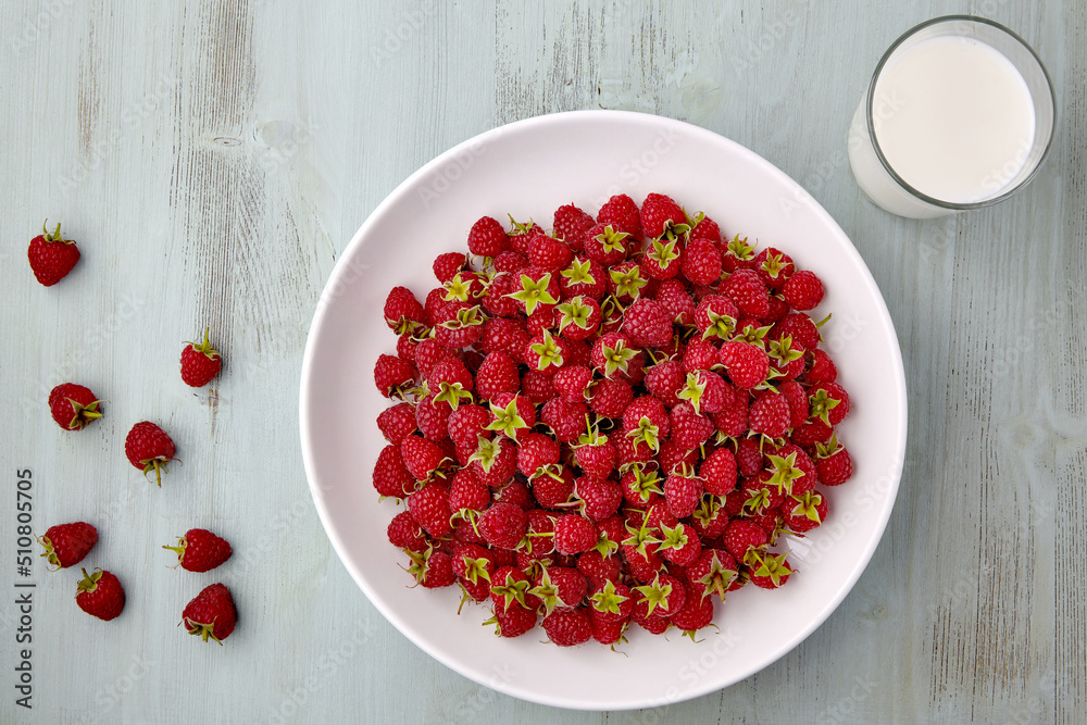 Ripe red raspberries with stalks are poured into a white ceramic plate and a glass of milk Ripe red raspberries with stalks are poured into a white ceramic plate and a glass of milk