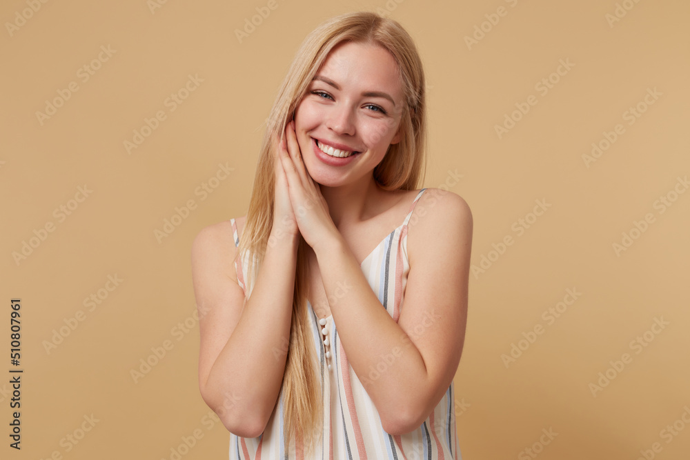 Studio portrait of young blonde woman posing over beige background smiling broadly and looking into camera, shows rest sign