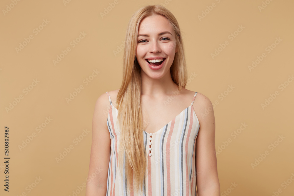 Studio portrait of young blonde woman posing over beige background smile broadly and looking into camera Studio portrait of young blonde woman posing over beige background smile broadly and looking into camera