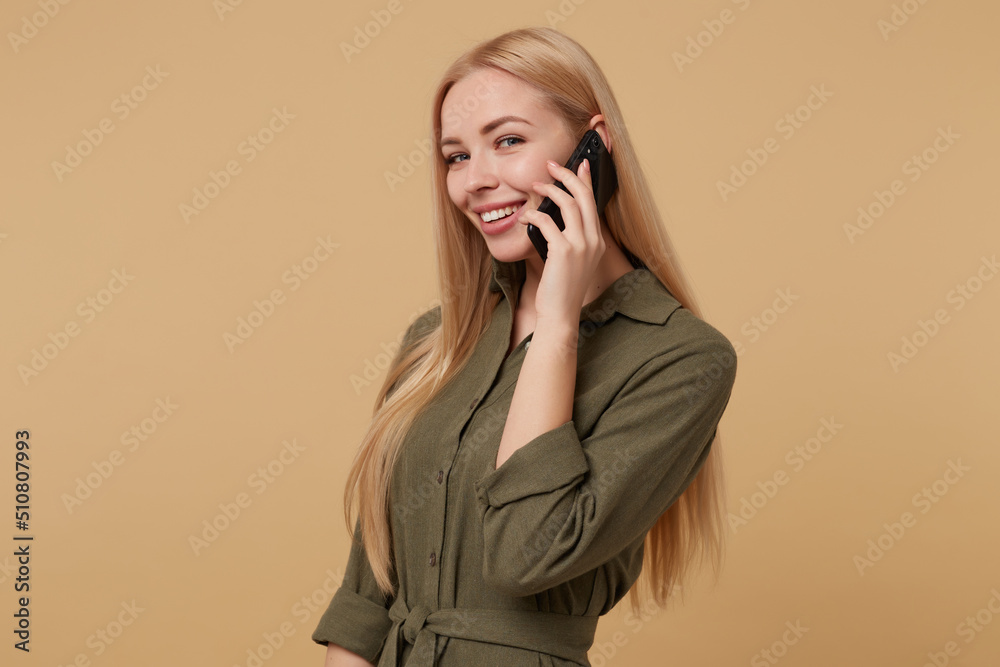 Studio portrait of young blonde woman posing over beige background smiles while talking on the phone