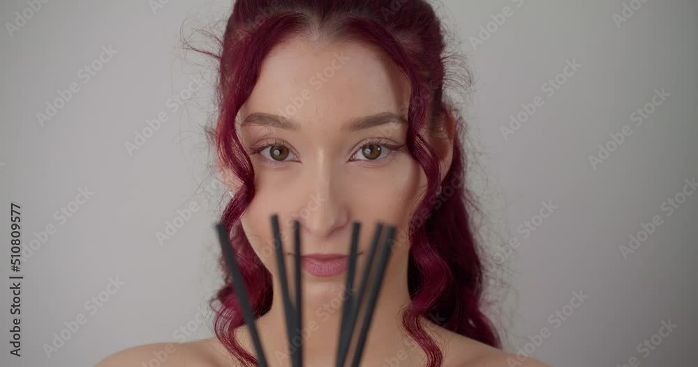 Pretty young indie hipster girl with pink purple hair holding incense in studio, looking into camera. White background, slow motion