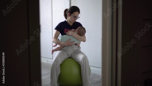 A young mother rocks her baby on a fitness ball. The girl puts the child to sleep at home.