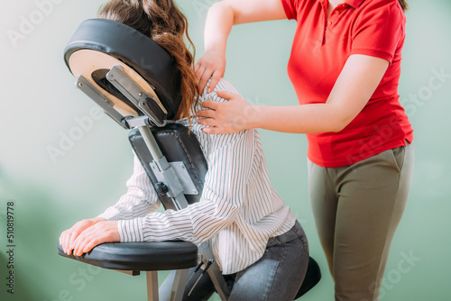 Female employee sitting on a portable massage chair in business office. Therapist massaging her shoulders, releasing muscle tension. photo