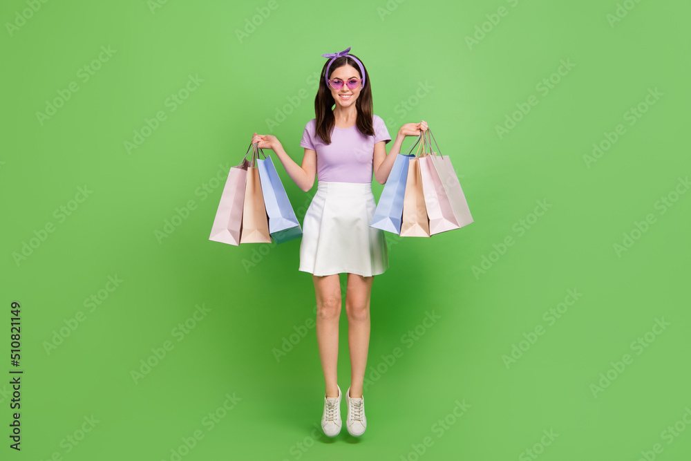 Full length photo of nice brunette lady hold bags jump wear glasses t-shirt skirt shoes isolated on green background Full length photo of nice brunette lady hold bags jump wear glasses t-shirt skirt shoes isolated on green background