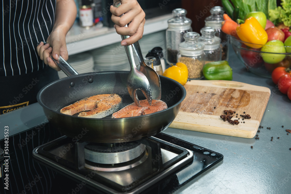 Woman chef salts raw salmon steak in a frying pan. Healthy seafood. Chef preparing salmon steak on stove. Cooking food concept. Woman chef salts raw salmon steak in a frying pan. Healthy seafood. Chef preparing salmon steak on stove. Cooking food concept.