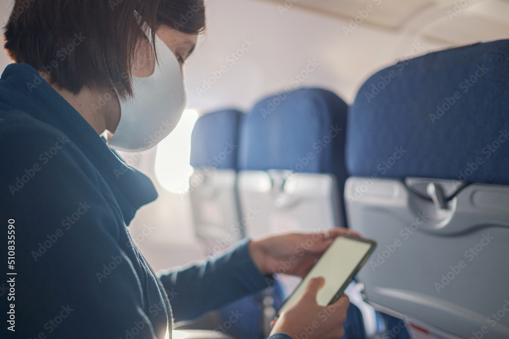 Young beautiful woman sitting at window of plane during the flight. new normal travel after covid-19 pandemic concept. shows smartphone monitor screen, mock-up