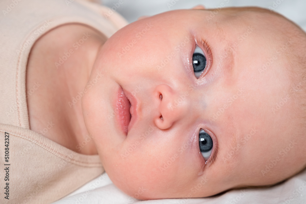 Portrait of beautiful newborn girl with big grey eyes lying on bed at home. Cute baby in beige bodysuit looks at camera in bedroom close view Portrait of beautiful newborn girl with big grey eyes lying on bed at home. Cute baby in beige bodysuit looks at camera in bedroom close view