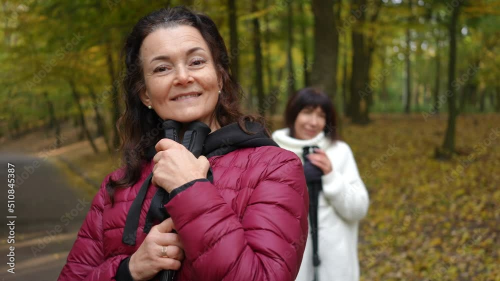 Positive smiling mature woman posing with ski poles and blurred friend at background. Portrait of happy confident Caucasian retiree looking at camera standing in autumn park. Exercising and enjoyment Positive smiling mature woman posing with ski poles and blurred friend at background. Portrait of happy confident Caucasian retiree looking at camera standing in autumn park. Exercising and enjoyment