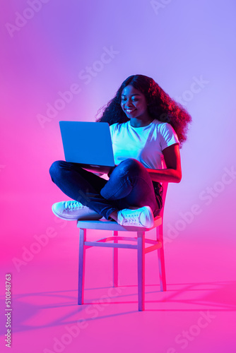 Happy young black woman using laptop pc for online work or studies, sitting on chair in neon light, full length