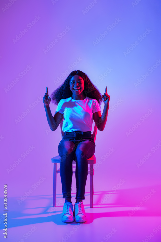 Cool promo. Young black lady sitting on chair, pointing upwards, offering empty space in neon light, mockup for ad