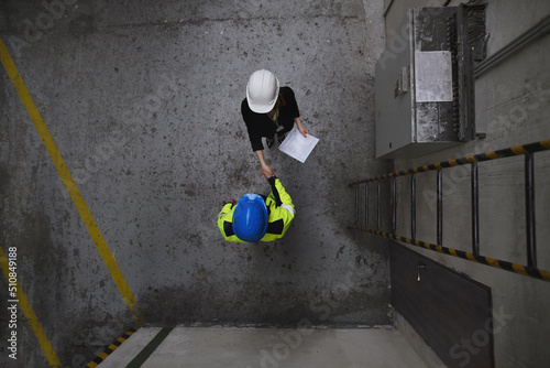 Top view of engineer and industrial worker in uniform shaking hands in large metal factory hall and talking.