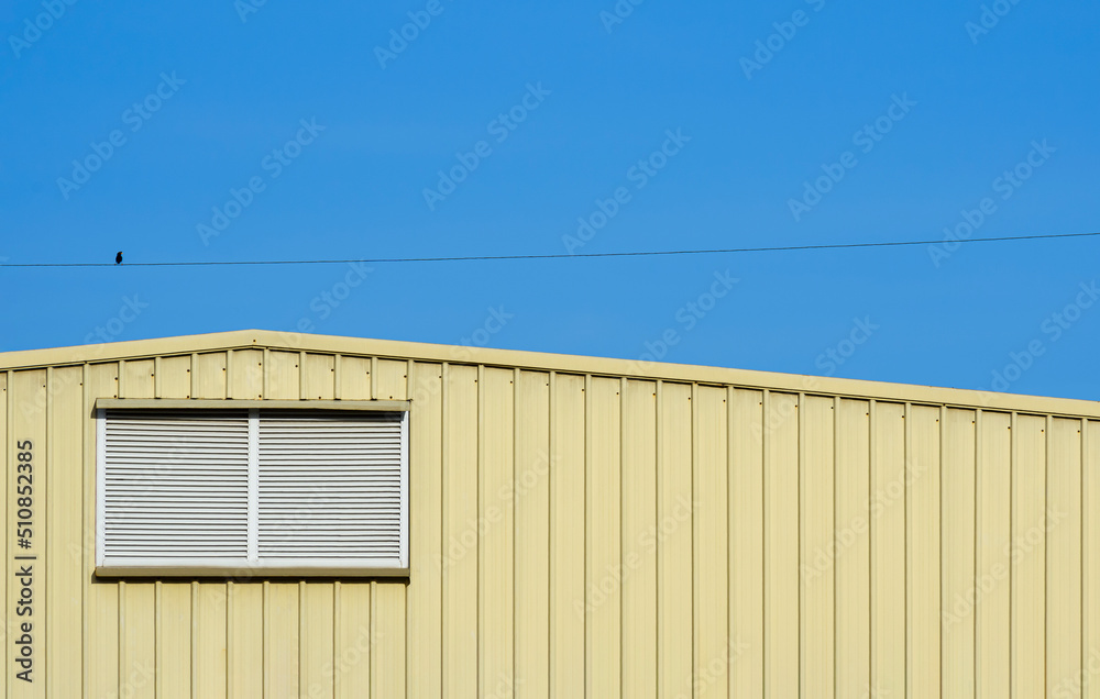 Aluminum louver on yellow corrugated steel warehouse wall with little bird perching on electric line against blue sky background Aluminum louver on yellow corrugated steel warehouse wall with little bird perching on electric line against blue sky background