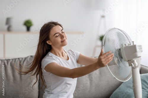 Satisfied pretty millennial caucasian female suffering from heat, sitting on sofa and enjoying cold air from fan